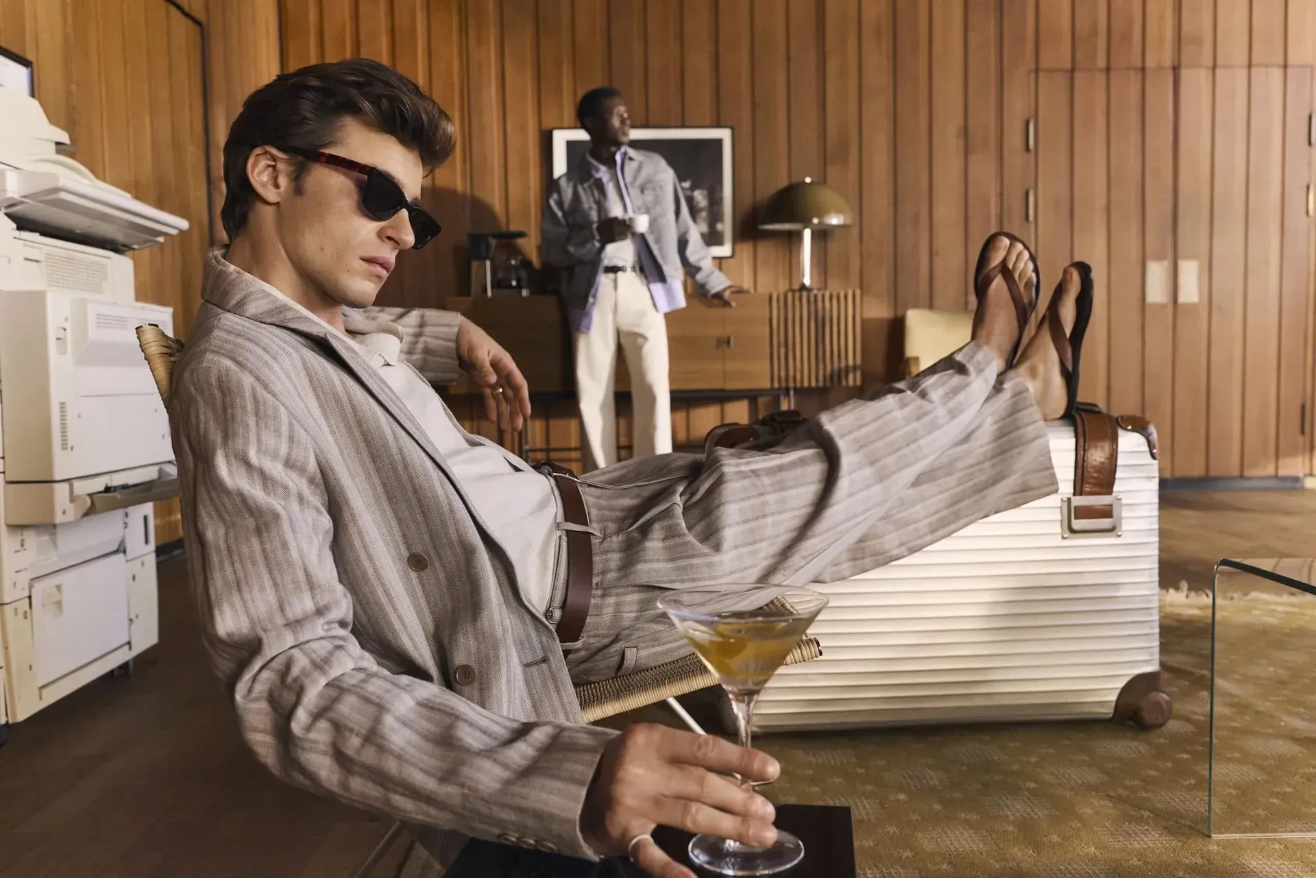 Campaign photograph from NN07: two men in a wood-panelled office, one reclining with his feet on a suitcase