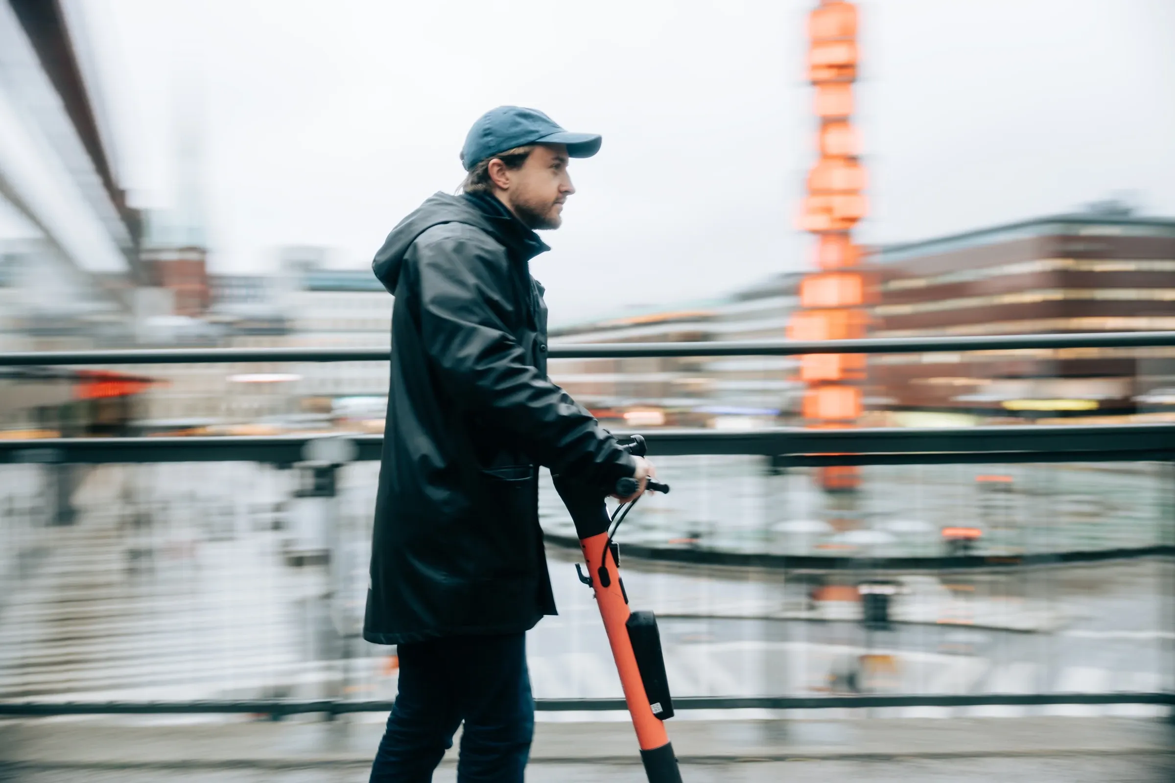 En man åker elsparkcykel längs ett räcke vid vattnet, staden i rörelseoskärpa bakom